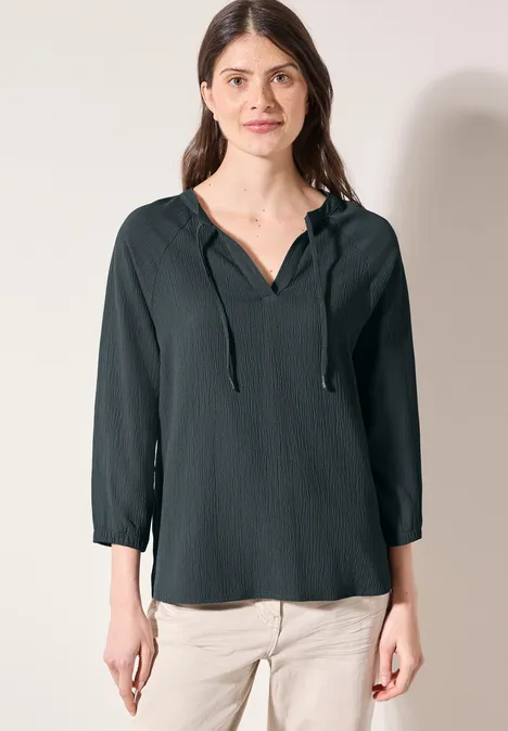 Frau in dunkelgrüner Bluse mit Bindeband und heller Hose.